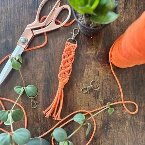 May include: Orange macrame keychain with a tassel, a pair of scissors, a spool of orange yarn, and two potted plants on a wooden surface.