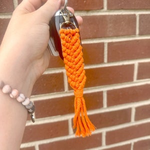 May include: Orange macrame keychain with a tassel. The keychain is attached to a silver key ring.