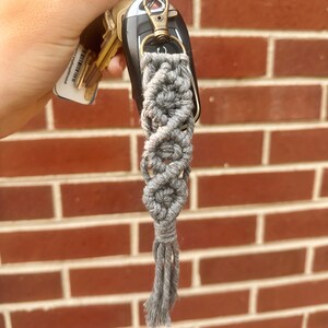 May include: A grey macrame keychain with a tassel hanging from a silver key ring. The keychain is attached to a set of keys, including a car key.