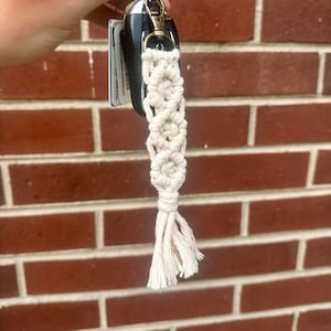 May include: A white macrame keychain with a key ring and a car key attached. The keychain is hanging from a hand.