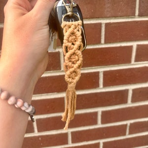 May include: A tan macrame keychain with a tassel hanging from a silver key ring. The keychain is attached to a set of keys.