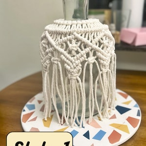 May include: A white macrame vase cover with fringe, sitting on a colorful tile coaster. The text "Style 1" is written in yellow on a white background.