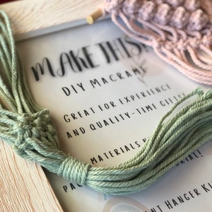 Puede incluir: Un marco de madera blanco con el texto "MAKE THIS DIY MACRAME GREAT FOR EXPERIENCE AND QUALITY-TIME GIFTS MATERIALS PAC UKIAL! PLANT HANGER KIT" impreso en un fondo blanco. Un cordón de macramé verde está envuelto alrededor del marco y un colgador de plantas de macramé rosa está en el fondo.