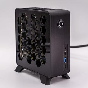 Peut inclure: Un boîtier d'ordinateur noir avec un motif en nid d'abeille sur le devant. Le boîtier rectangulaire a des bords arrondis et est soutenu par de petits pieds. Il comprend un bouton d'alimentation, des ports USB et une prise casque. Mesure environ 20 cm de haut.