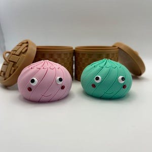 May include: Two rounded, swirled dumpling figures with googly eyes and small red cheeks, one light pink and one mint green. Each figure sits in front of a small, brown, cylindrical container with a woven texture and a matching removable lid.
