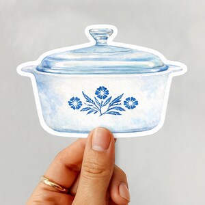 May include: A sticker of a blue and white casserole dish with a lid. The dish features a floral design in blue. The sticker is held by a hand with a gold ring.