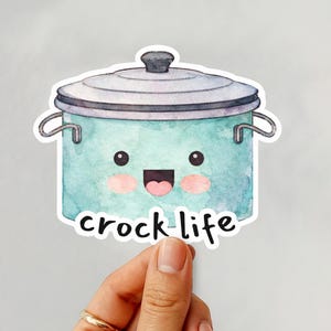 May include: A watercolor sticker featuring a smiling teal-colored slow cooker with a lid. The sticker has the words "crock life" in a handwritten font. The sticker is held up by a hand with a gold ring.