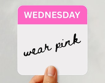 May include: A hand holding a white card with a pink top section that reads "WEDNESDAY". Below, in black cursive, it says "wear pink". A gold ring is on the person's finger.