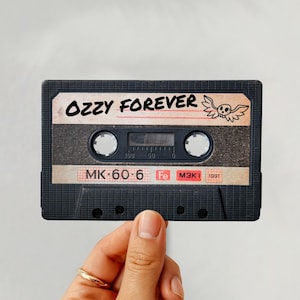 May include: A black cassette tape with the text "OZZY FOREVER" and a skull graphic. The tape has a label with the text "MK-60-6". The cassette is held by a hand with a gold ring.