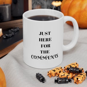 May include: White ceramic coffee mug with the text "JUST HERE FOR THE COMMENTS" printed in black. The mug is filled with coffee and steam is rising from the top. The mug is sitting on a gray fabric surface with orange and black candy.