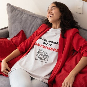 May include: A woman wearing a white t-shirt with the text "DIGITAL NOMAD DO IT ANYWHERE" and a red blazer, sitting on a gray couch.