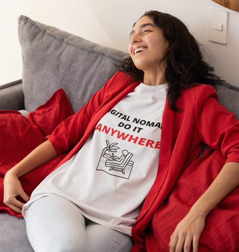 May include: A woman wearing a white t-shirt with the text "DIGITAL NOMAD DO IT ANYWHERE" and a red blazer, sitting on a gray couch.