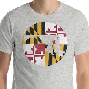Op de afbeelding: Een grijs gemêleerd t-shirt met korte mouwen en een ronde afbeelding op de borst. De afbeelding toont het patroon van de vlag van de staat Maryland met een witte silhouet van de staat Maryland in het midden van de cirkel.