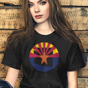 Puede incluir: Una camiseta negra con un gráfico circular desgastado que presenta el estado de Arizona en rojo, amarillo y azul. El gráfico está centrado en la camiseta.