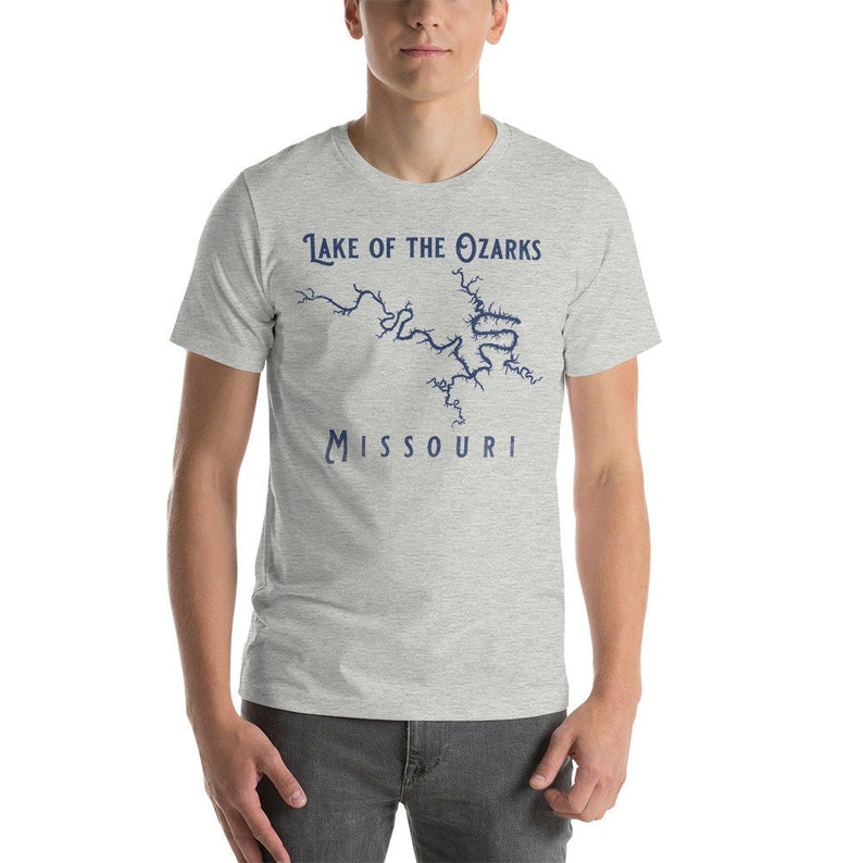 May include: A light gray t-shirt with the text "Lake of the Ozarks Missouri" in navy blue. The design includes a navy blue outline of the lake's shape. The shirt is short-sleeved.