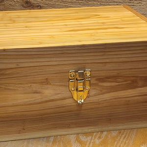 May include: A rectangular wooden storage box featuring a natural light brown wood grain finish. The box is constructed with visible finger joints at the corners and includes a shiny, silver-toned metal latch centered on the front panel.