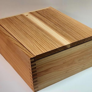 May include: A wooden box with a hinged lid. The box is made of light-colored wood with a natural grain pattern. The lid is closed and the box is sitting on a white surface.