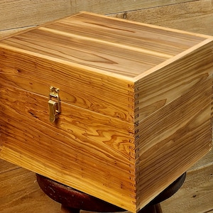 May include: A handcrafted wooden box with a natural wood grain finish. The box features a brass-colored clasp and is constructed with dovetail joints. The box is sitting on a small wooden stool.