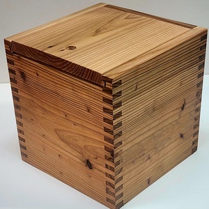 May include: A handcrafted wooden box with a lid. The box is constructed from light brown wood with visible grain and features dovetail joints at the corners. The lid has a small circular hole.