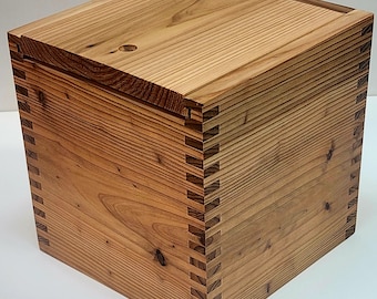 May include: A handcrafted wooden box with a lid. The box is constructed from light brown wood with visible grain and features dovetail joints at the corners. The lid has a small circular hole.