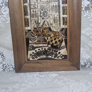 Puede incluir: Una pieza de arte mural rectangular con un marco de madera marrón que rodea un azulejo de cerámica texturizado. El azulejo representa una escena en marrón oscuro y crema de una fachada de edificio con un rosetón, un frutero y un paño a cuadros.