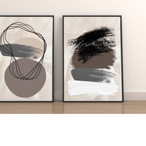 May include: Two framed abstract art prints with a beige background. The print on the left features a brown circle with black lines and a gray brushstroke. The print on the right features a black brushstroke, a brown brushstroke, a gray brushstroke, and a white brushstroke.