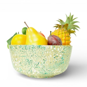 Puede incluir: Un cuenco decorativo lleno de fruta fresca, incluyendo una piña, una ciruela, una pera y un limón. El cuenco tiene un patrón moteado verde y crema. La fruta está dispuesta sobre un fondo blanco.