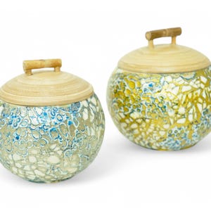 May include: Two round decorative storage containers with lids. The containers are made of bamboo and have a mosaic-like pattern. One container is blue and white, and the other is gold and white.
