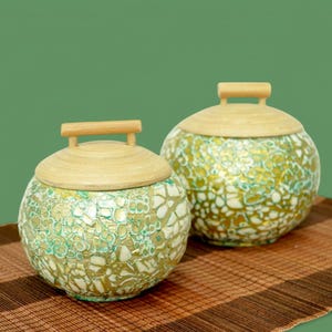 May include: Two round, decorative storage containers with wooden lids and handles. The containers are made of a mosaic-like material with green, gold, and white accents. The containers are sitting on a brown woven mat.
