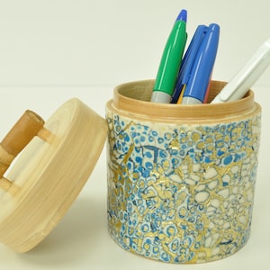 May include: A wooden pencil holder with a blue and gold mosaic design. The lid has a wooden handle. There are four pens and a marker inside the holder.