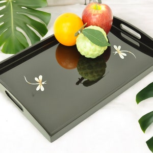 May include: A black lacquer serving tray with two dragonfly designs. The tray has a raised edge and two handles. There are three pieces of fruit on the tray: a red apple, a yellow orange, and a green pomelo.