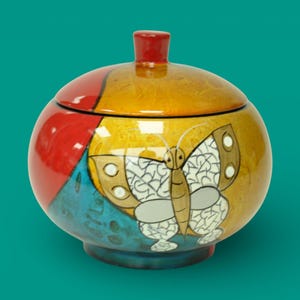 Op de afbeelding: Een ronde keramische pot met deksel en vlinderontwerp. De pot is beschilderd met een mix van kleuren, waaronder rood, geel, blauw en wit. Het deksel heeft een kleine rode knop.
