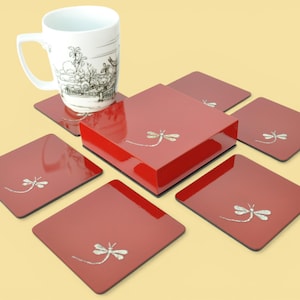 May include: A set of red square coasters with a glossy finish, each adorned with a silver dragonfly design. A matching red square box sits in the center, and a white mug with a black and white illustration rests on top of the coasters.