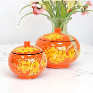 May include: Two orange, round decorative boxes with lids, adorned with yellow floral patterns. The boxes have a glossy finish and are of different sizes. A vase with flowers is in the background.