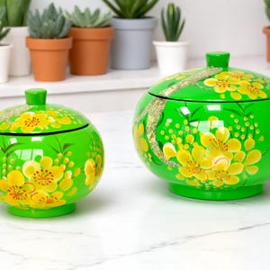 Puede incluir: Dos tarros de cerámica verdes con tapas, adornados con diseños florales amarillos y detalles dorados. Los tarros son redondos con un pequeño pomo en la parte superior. El fondo presenta plantas en macetas en un estante.