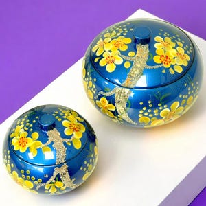 Op de afbeelding: Twee blauwe, ronde decoratieve dozen met deksels. De dozen zijn versierd met gele bloempatronen en een zilverkleurig hagedisachtig ontwerp. Ze staan op een wit oppervlak met een paarse achtergrond.