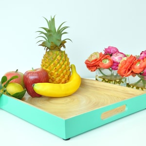 Può includere: Un vassoio rettangolare in bambù con un bordo verde acqua brillante, contenente un ananas, mele rosse, una banana e un limone. Due vasi di vetro con fiori colorati a destra.