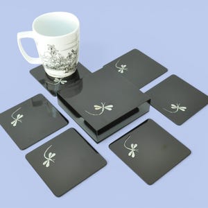May include: A set of six black square coasters with a matching box, each adorned with a delicate silver dragonfly design. A white mug with a black illustration sits atop the coasters, all on a light blue surface.