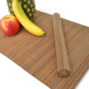Puede incluir: Un mantel individual rectangular de color marrón tejido con una superficie texturizada. Un mantel está extendido, mientras que otro está enrollado en forma de cilindro. Una manzana roja, un plátano amarillo y una piña están sobre el mantel.