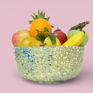 Puede incluir: Un cuenco decorativo lleno de varias frutas, incluyendo una naranja, una manzana, una pera, un plátano y una piña. El cuenco tiene un patrón moteado en tonos de amarillo, azul y blanco. El fondo es rosa liso.