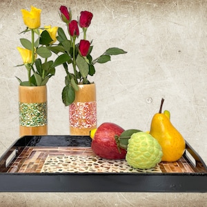 May include: A black tray with a mosaic design, featuring yellow roses in a green and gold vase, red roses in an orange and white vase, a red apple, a green cherimoya, and a yellow pear.