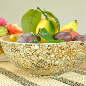 May include: A gold and white ceramic bowl filled with fruit, including lemons, plums, and bananas. The bowl has a textured, mosaic-like design.