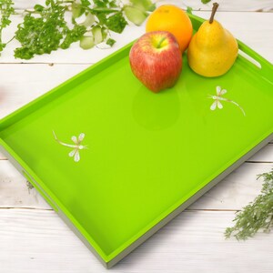 May include: A green serving tray with a raised edge and two dragonfly designs. The tray is on a white wooden surface with fruit on top, including a red apple, a yellow pear, and an orange.