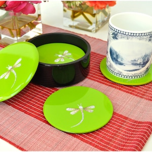 Puede incluir: Juego de cuatro posavasos verdes con un diseño de libélula. Los posavasos son redondos y tienen un acabado brillante. Los posavasos están en una caja negra con tapa.