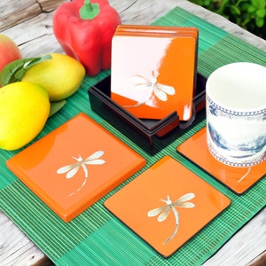 May include: Set of four orange square coasters with a white dragonfly design. The coasters are in a black box and are sitting on a green bamboo mat. A white mug with a blue and white design is sitting on one of the coasters.