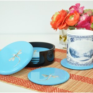 May include: Set of six blue coasters with a dragonfly design. The coasters are in a black holder and are on a woven placemat.