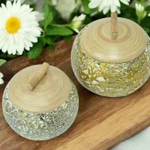 May include: Two round, decorative boxes with lids made of natural wood and a mosaic design. The boxes are on a wooden surface and have a gold and white mosaic pattern.