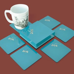 May include: A white ceramic mug with a black and white illustration sits atop a set of six square, turquoise coasters. Each coaster features a silver dragonfly design. The coasters are arranged around a turquoise box.