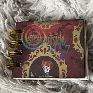 May include: A small, burgundy-colored spiral bound notebook with a gold and black ornate design. The cover features a skeleton with red hair and a floral headpiece. The text "Outside the Lines" is visible on the cover.