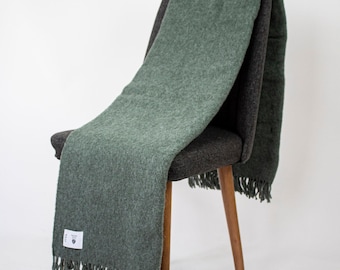 May include: A sage green throw blanket with a textured, woven appearance and fringe detailing along the bottom edge. A small white rectangular label with black text is attached to the lower corner of the blanket, which is draped over a dark chair.